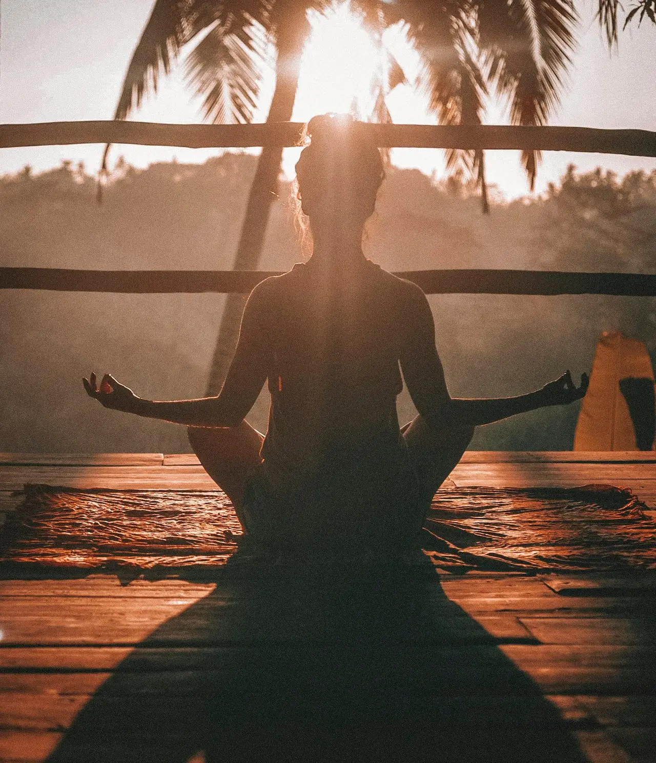 A person sitting cross-legged in a meditation pose on a wooden deck at sunrise